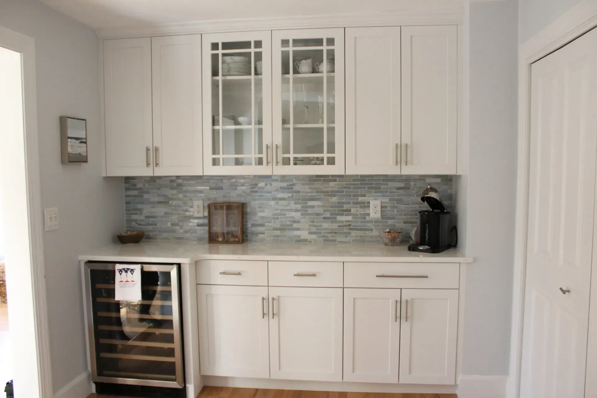 White kitchen cabinets with glass-front uppers, blue mosaic backsplash, wine fridge, and coffee maker.