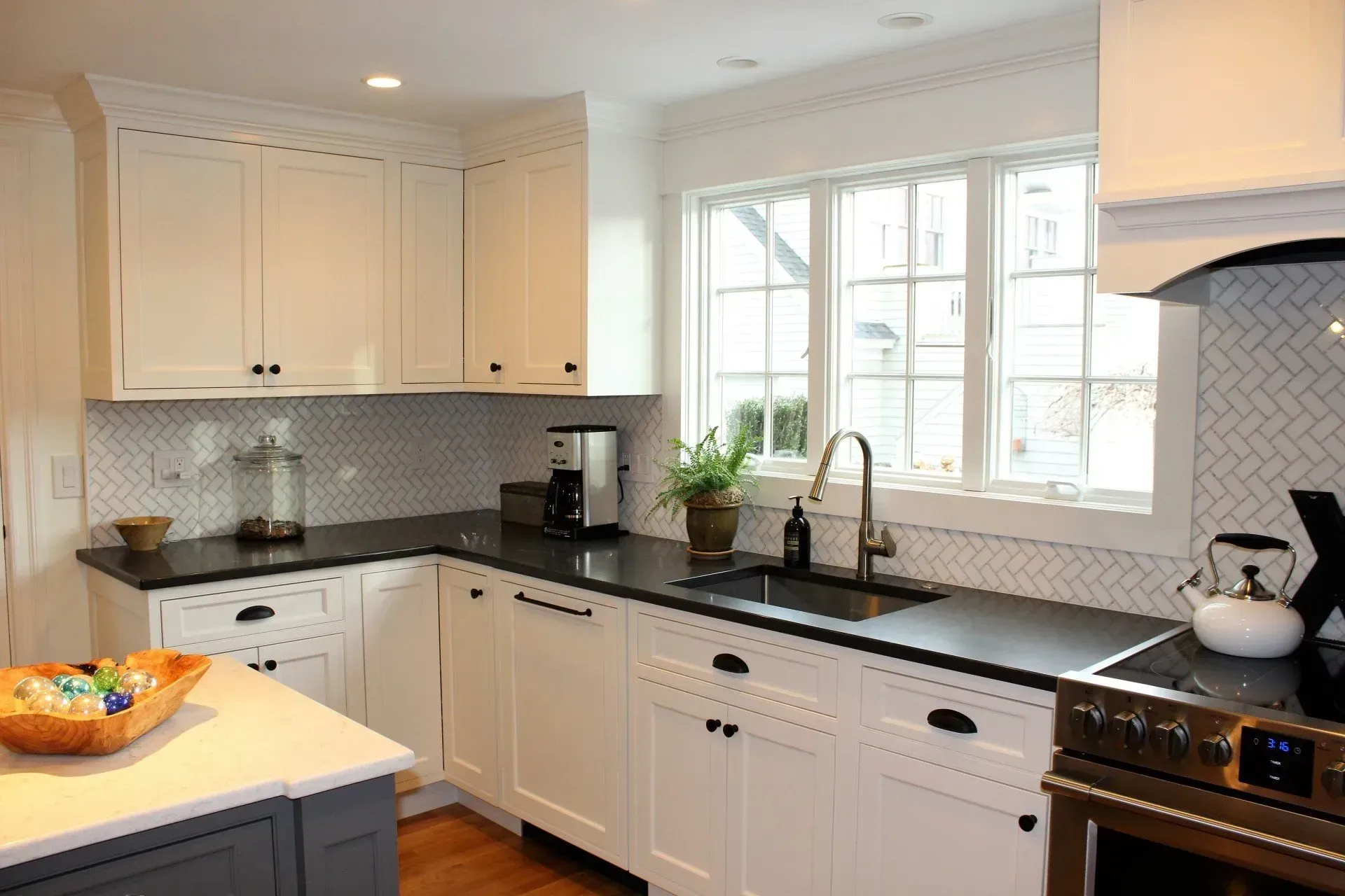 White kitchen with black countertops, cabinets, and appliances. Herringbone tile backsplash and window.