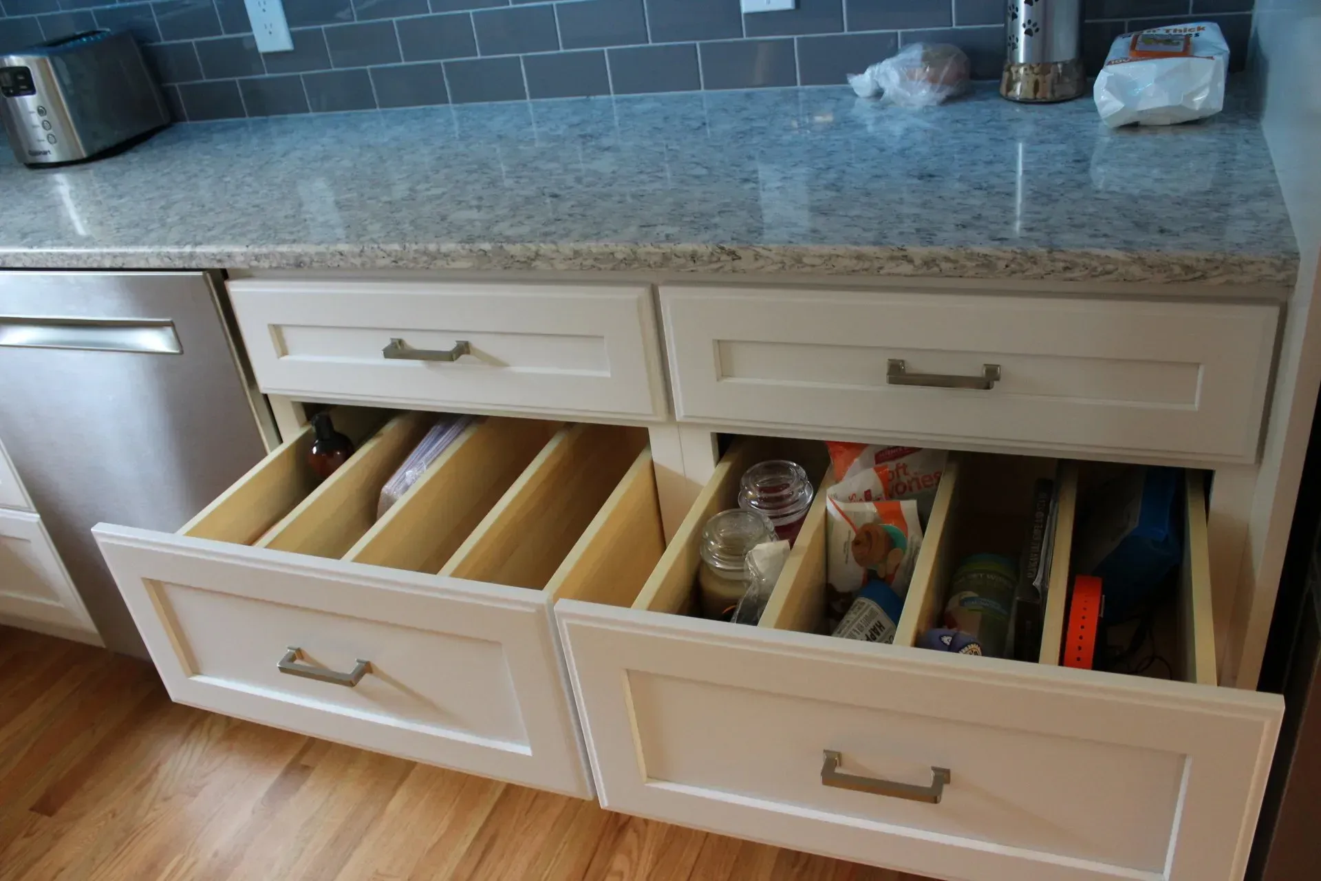 Kitchen drawers with dividers, containing utensils and supplies. White cabinets, granite countertop, wooden floor.