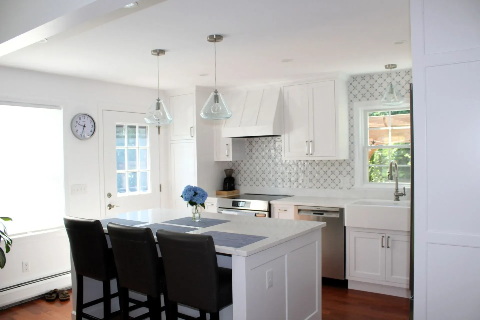 White kitchen with island and three bar stools, pendant lights, and a small blue flower arrangement.