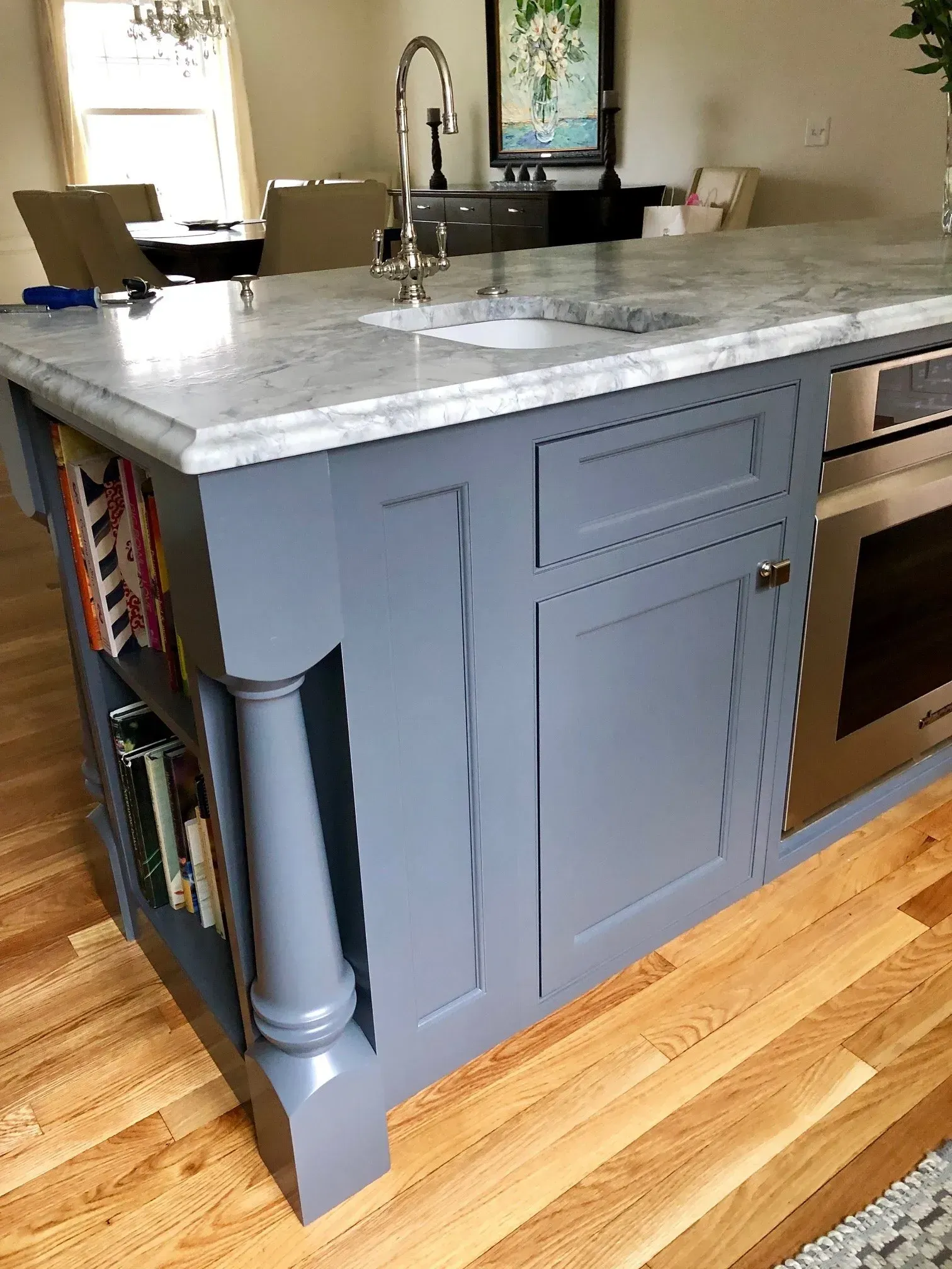 Gray kitchen island with marble countertop, built-in microwave, and bookshelf.