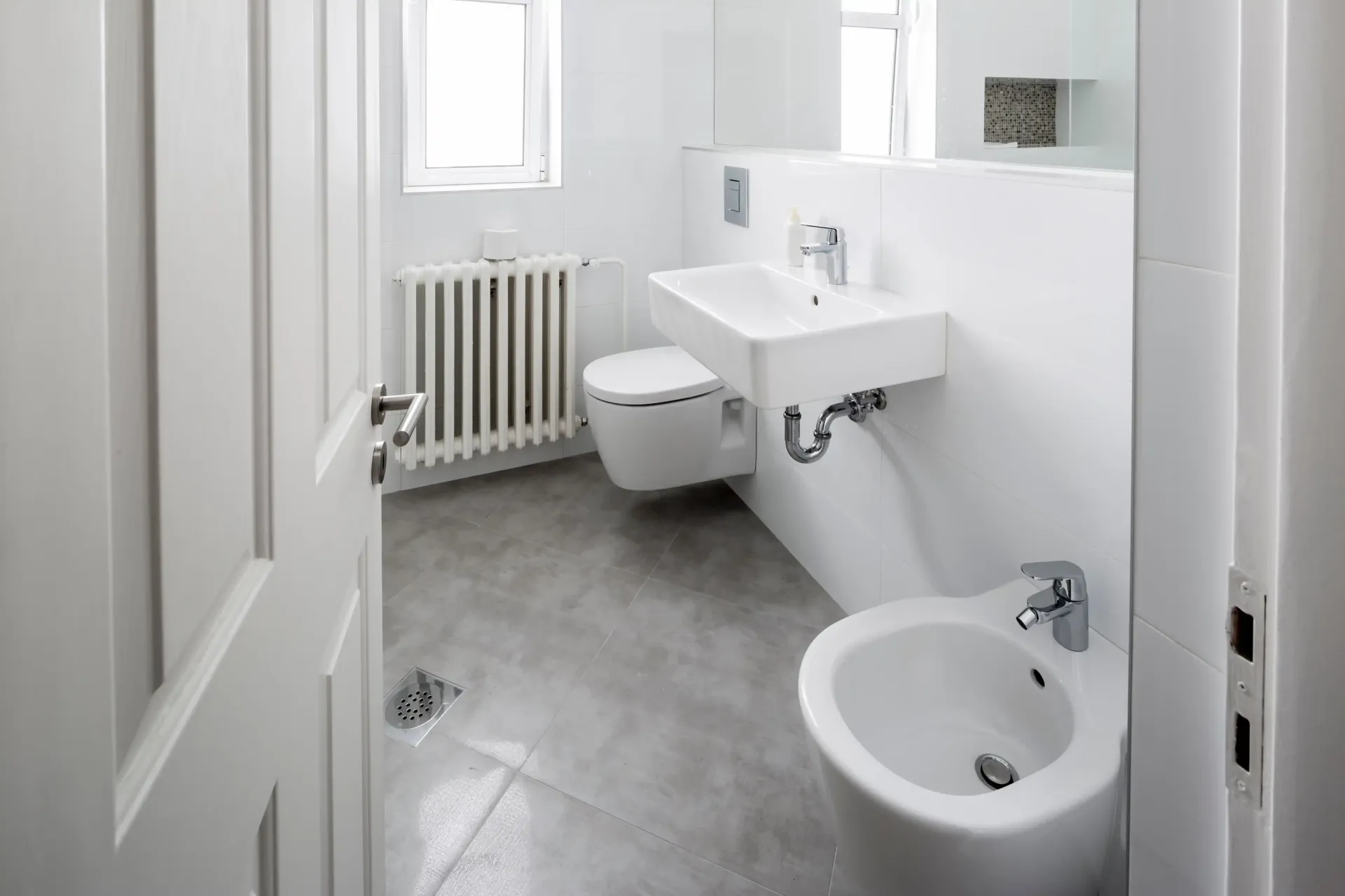 Brandon Handyman: A modern, minimalist white bathroom with a sink, toilet, and bidet, gray floor, and a radiator.