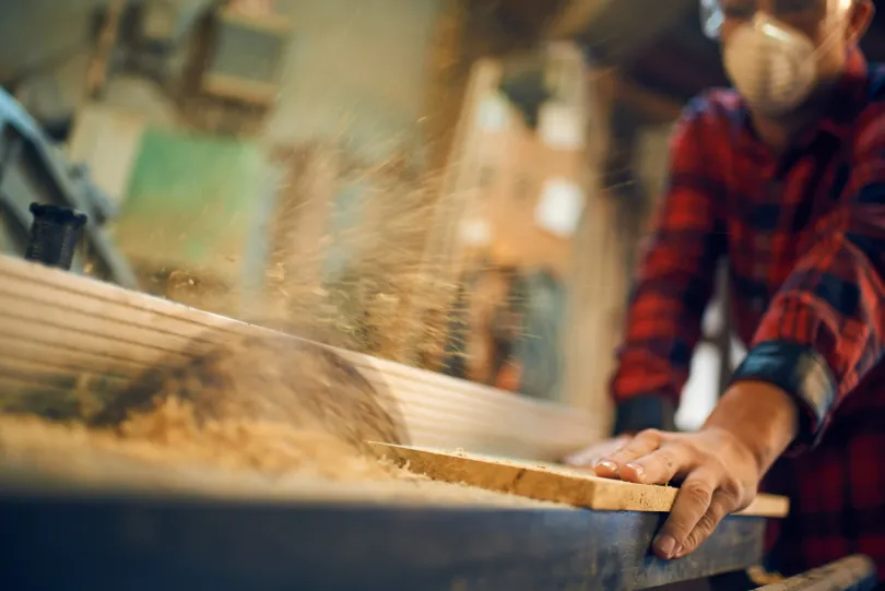 Brandon Handyman: Woodworker in red plaid shirt, cutting a wooden board with a saw, sawdust flying.
