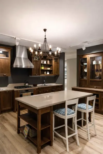 Brandon Handyman: Modern kitchen with a wooden dining table and black chairs, under a row of pendant lights. Dark grey cabinets and stainless steel appliances are visible.