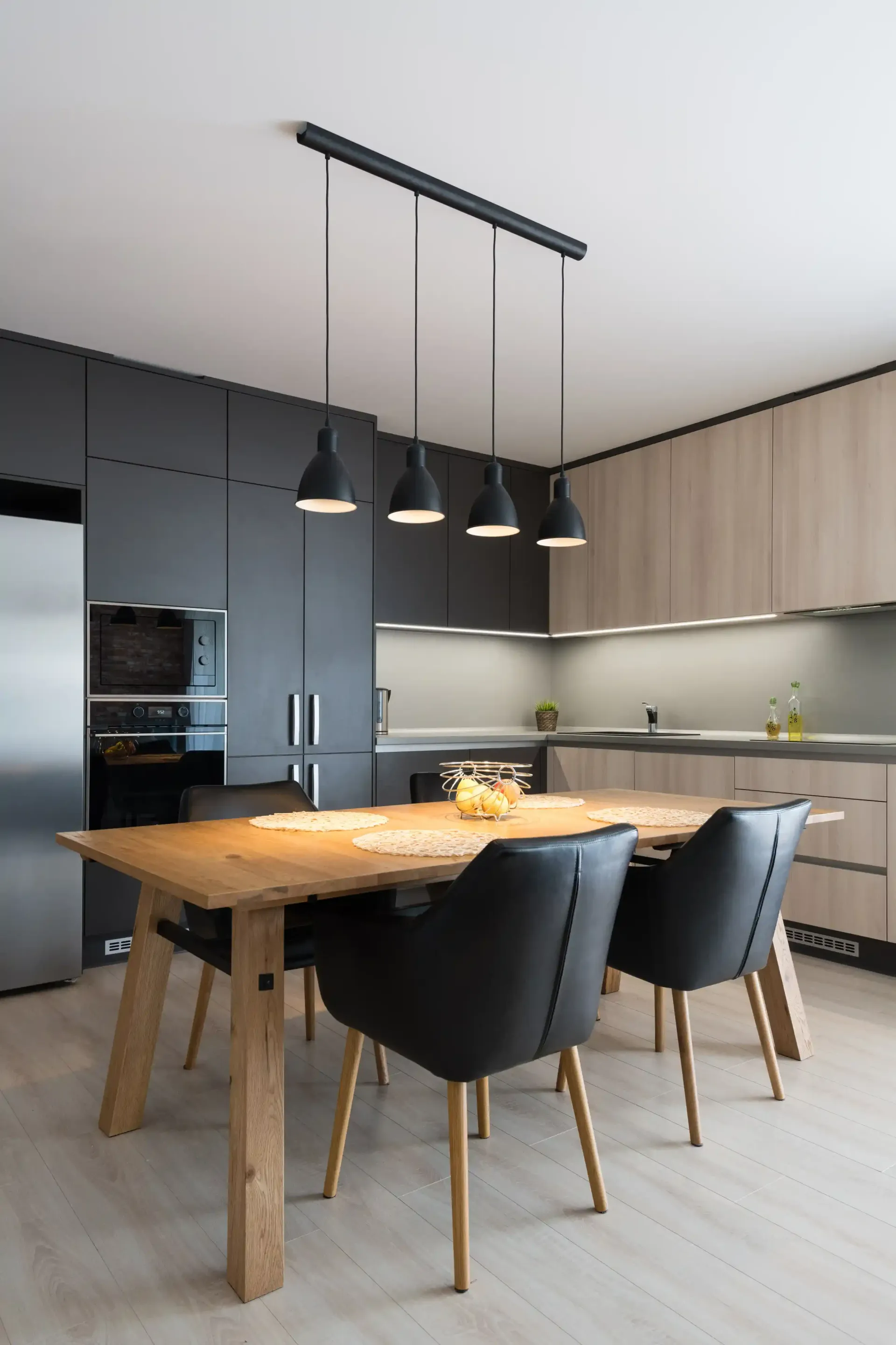 Brandon Handyman: Modern kitchen with a wooden dining table and black chairs, under a row of pendant lights. Dark grey cabinets and stainless steel appliances are visible.
