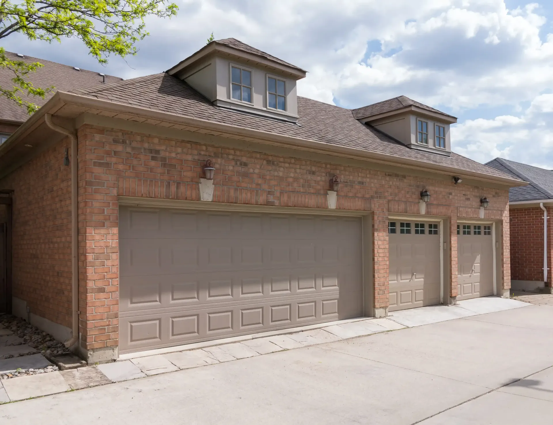 Brandon Handyman: Three-car brick garage with brown doors, two dormers, and light-colored trim against a cloudy sky.