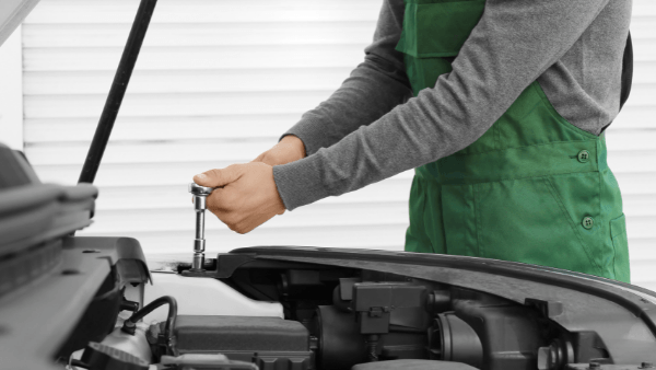 a mechanic taking out mobile mechanic services in Hastings. He is wearing a green apron and using a chrome tool under the bonnet of a car.