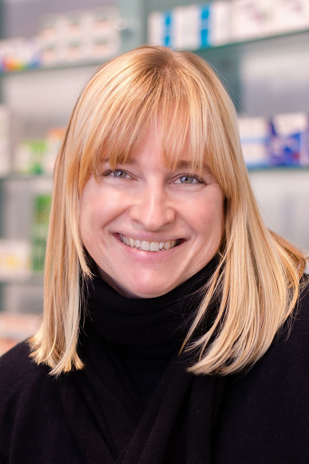 Eine Frau in einem schwarzen Rollkragenpullover lächelt in einer Apotheke.