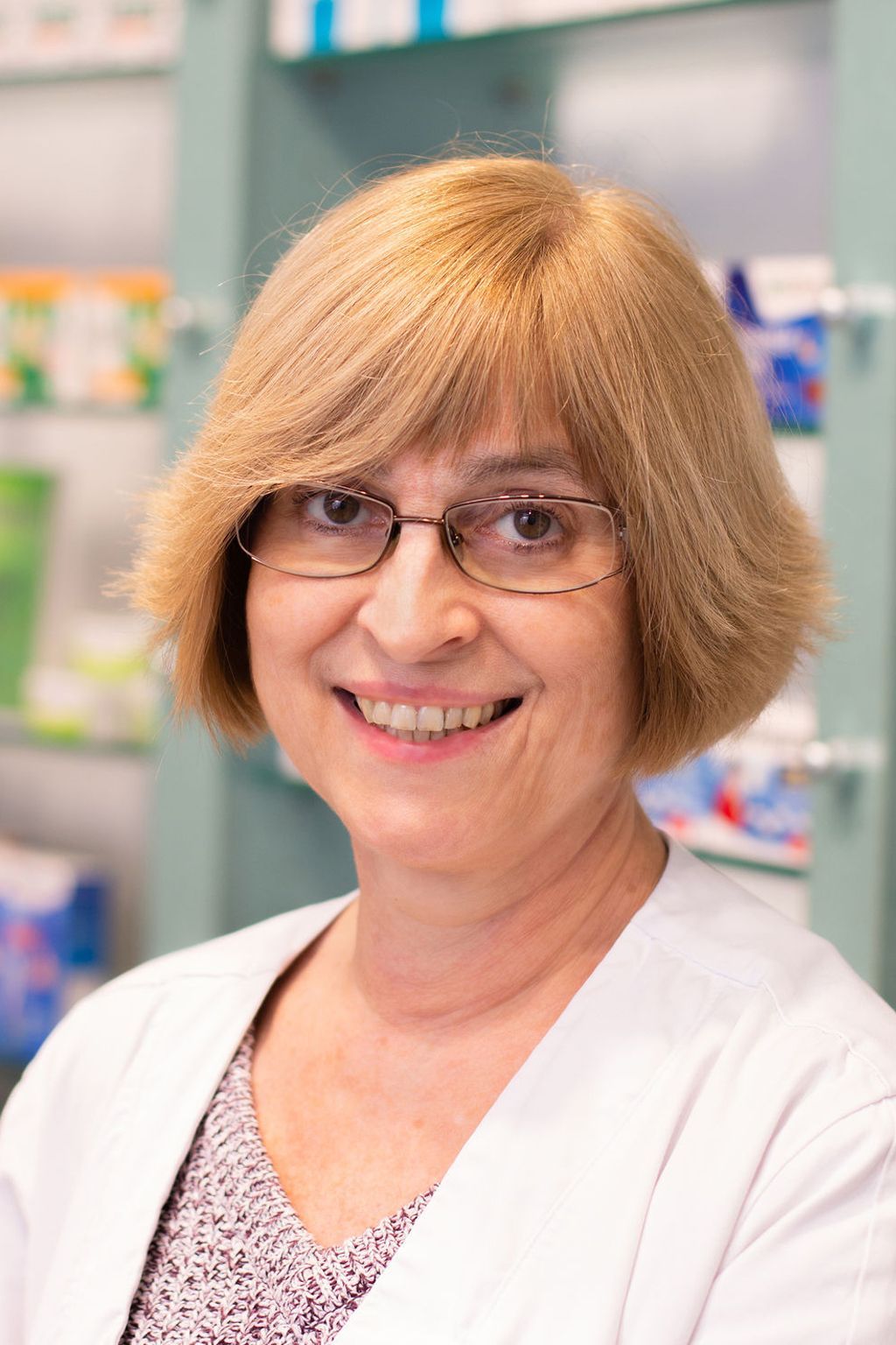 Eine Frau mit Brille und weißem Kittel lächelt in einer Apotheke.