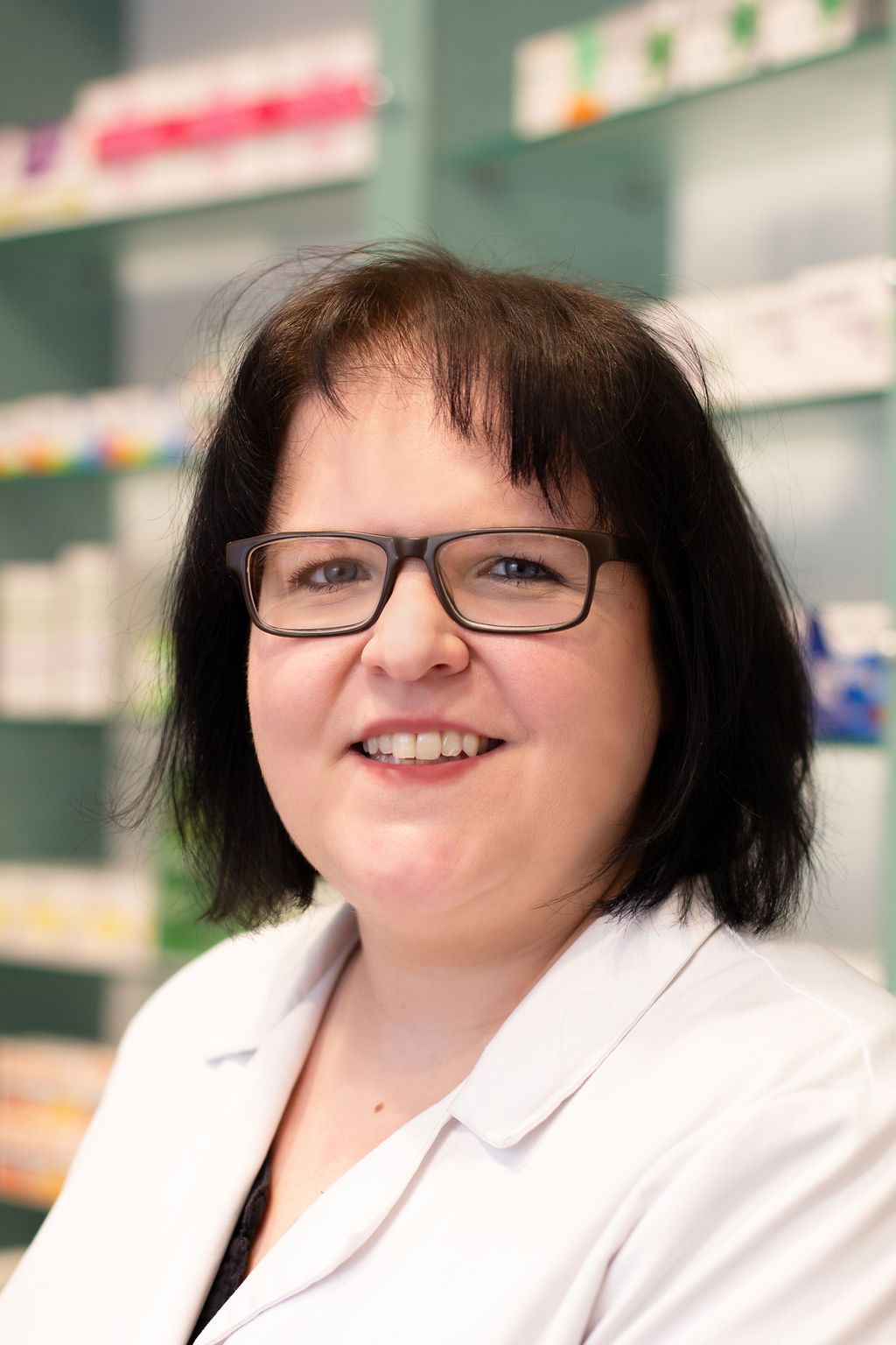 Eine Frau mit Brille und weißem Kittel lächelt in einer Apotheke.