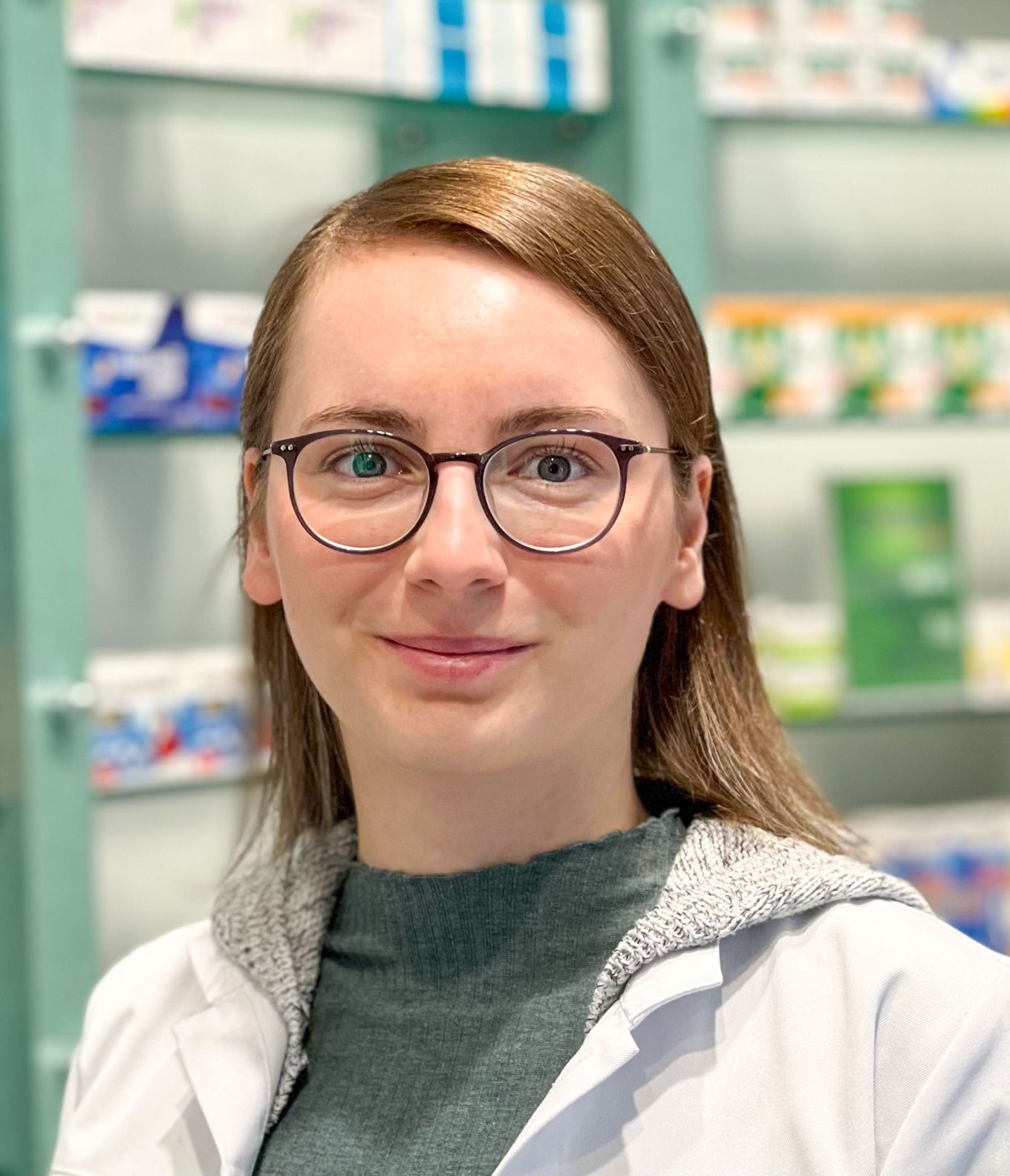 Eine Frau mit Brille und Laborkittel steht vor einer Apotheke.