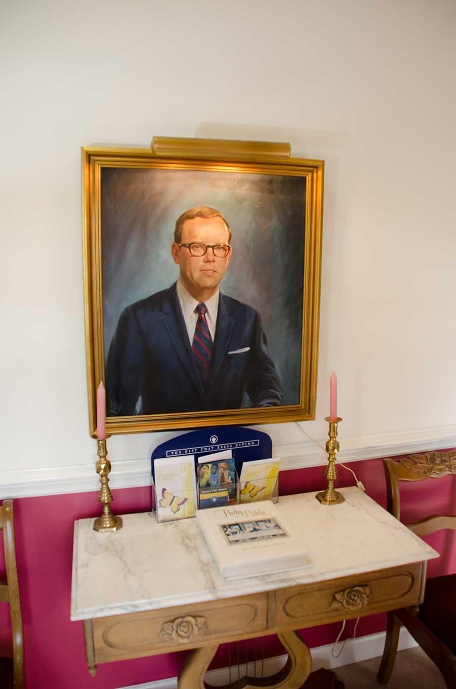 Portrait of a man in a suit, above a table with items. Candles flank the table against a pink wall.
