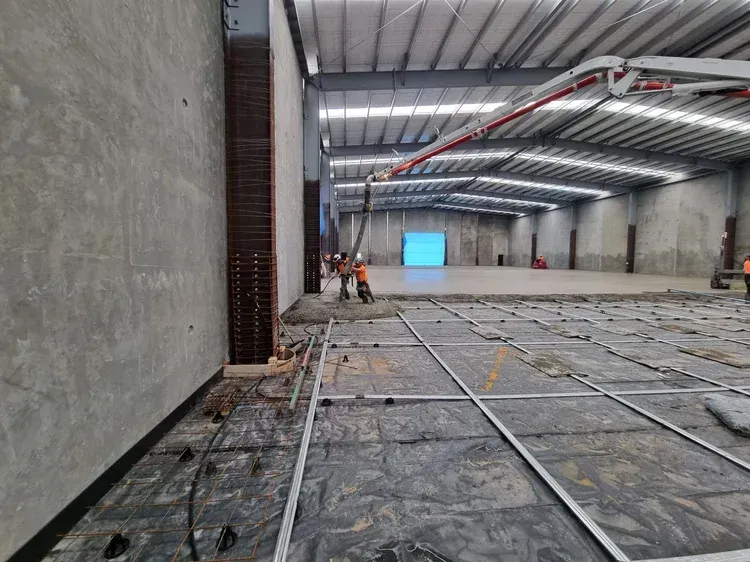 Workers pouring concrete floor in a large warehouse. A concrete pump arm extends, delivering the mixture.