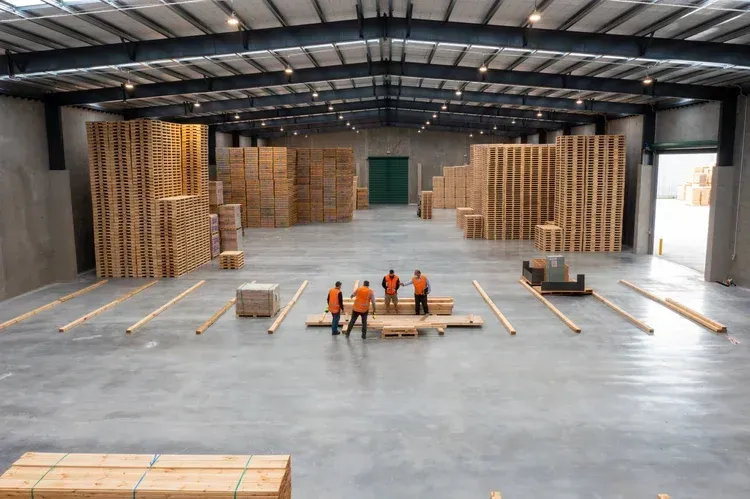 Four workers in vests in a large warehouse surrounded by stacks of wooden pallets.