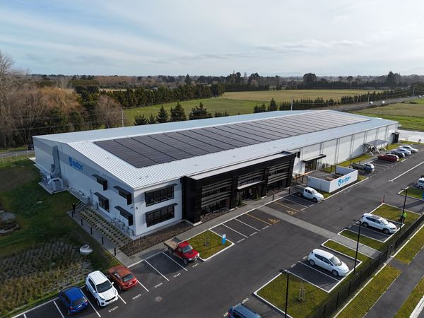 Aerial view of a modern industrial building with solar panels, cars parked in a lot, and surrounding greenery.