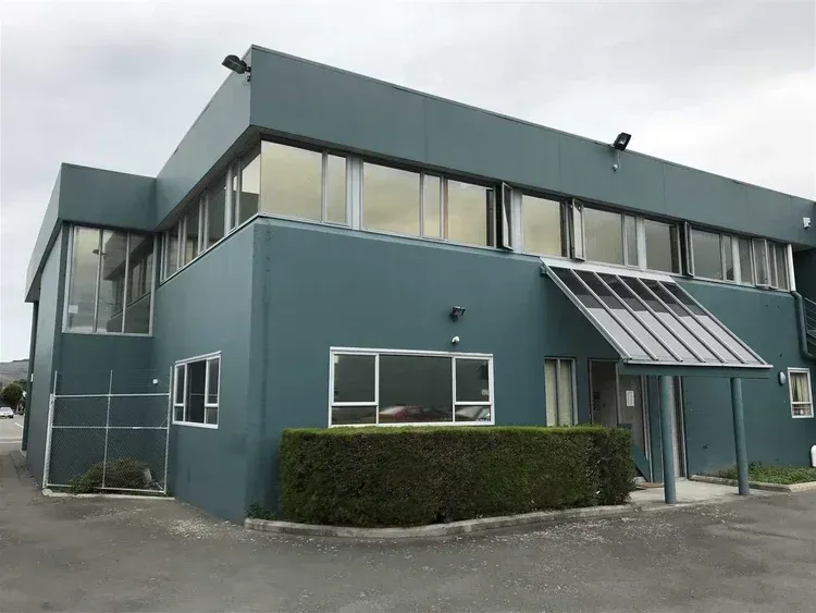 Two-story teal building with many windows, glass awning, and short green hedge in front. Cloudy sky overhead.