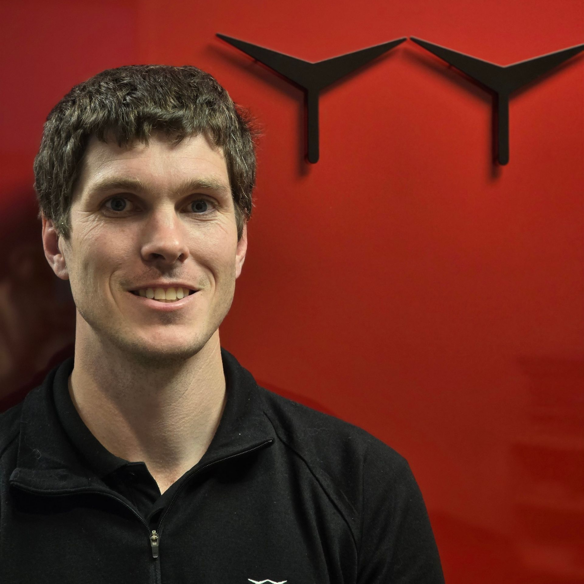 Man smiling in front of a red wall with black geometric logo, wearing a black jacket.