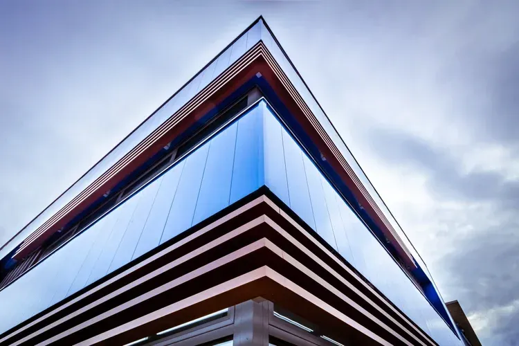 Angular modern building with blue glass facade and striped, brown overhang against a cloudy sky.