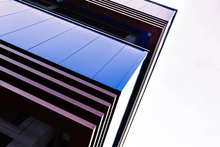 Looking up at modern building's geometric facade with blue panels and reddish-brown bands, against a bright sky.