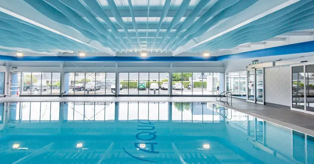 Indoor swimming pool with blue water, wavy ceiling, and large windows overlooking parking lot.