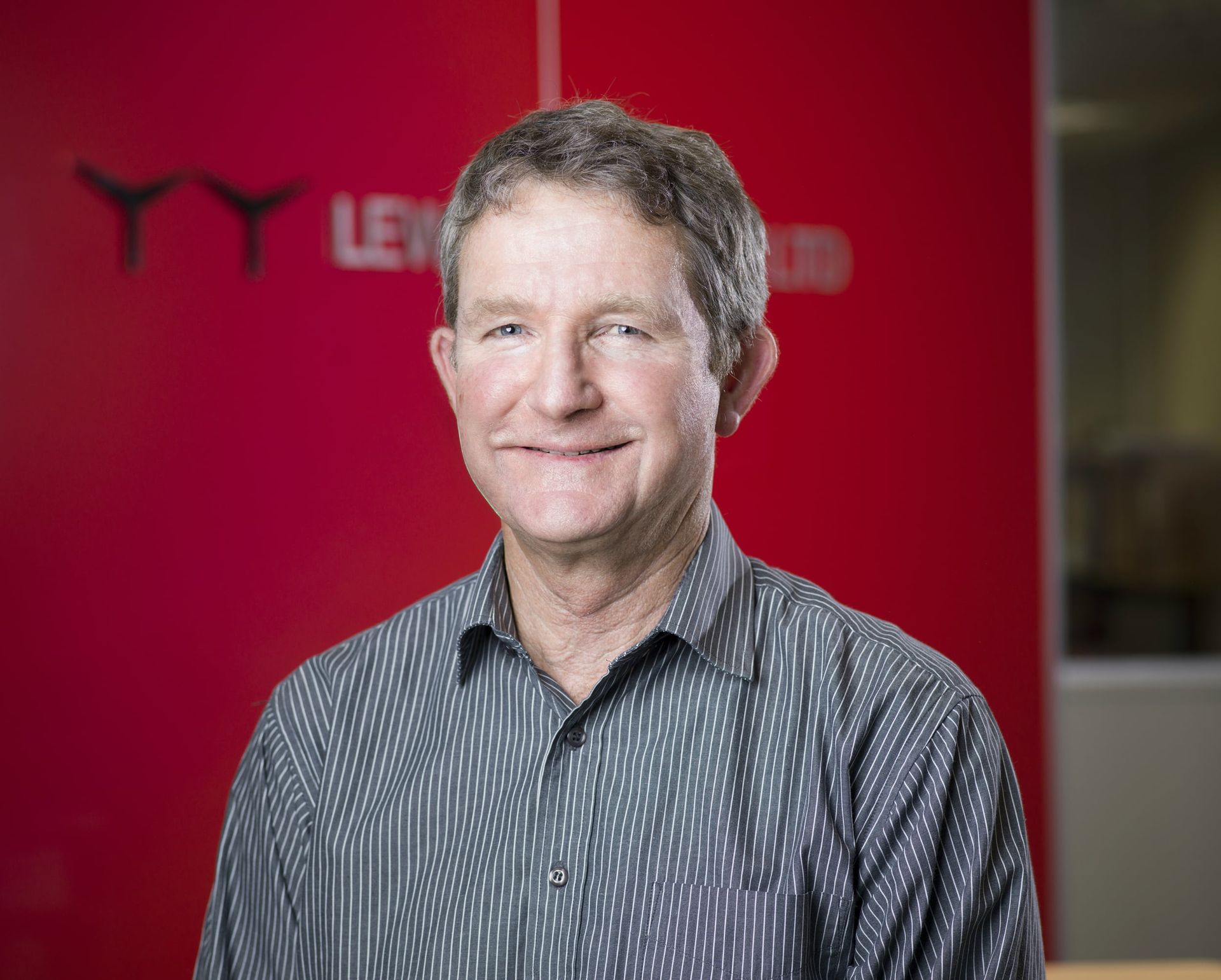 Man with graying hair smiles, wearing striped shirt in front of a red wall with a logo.