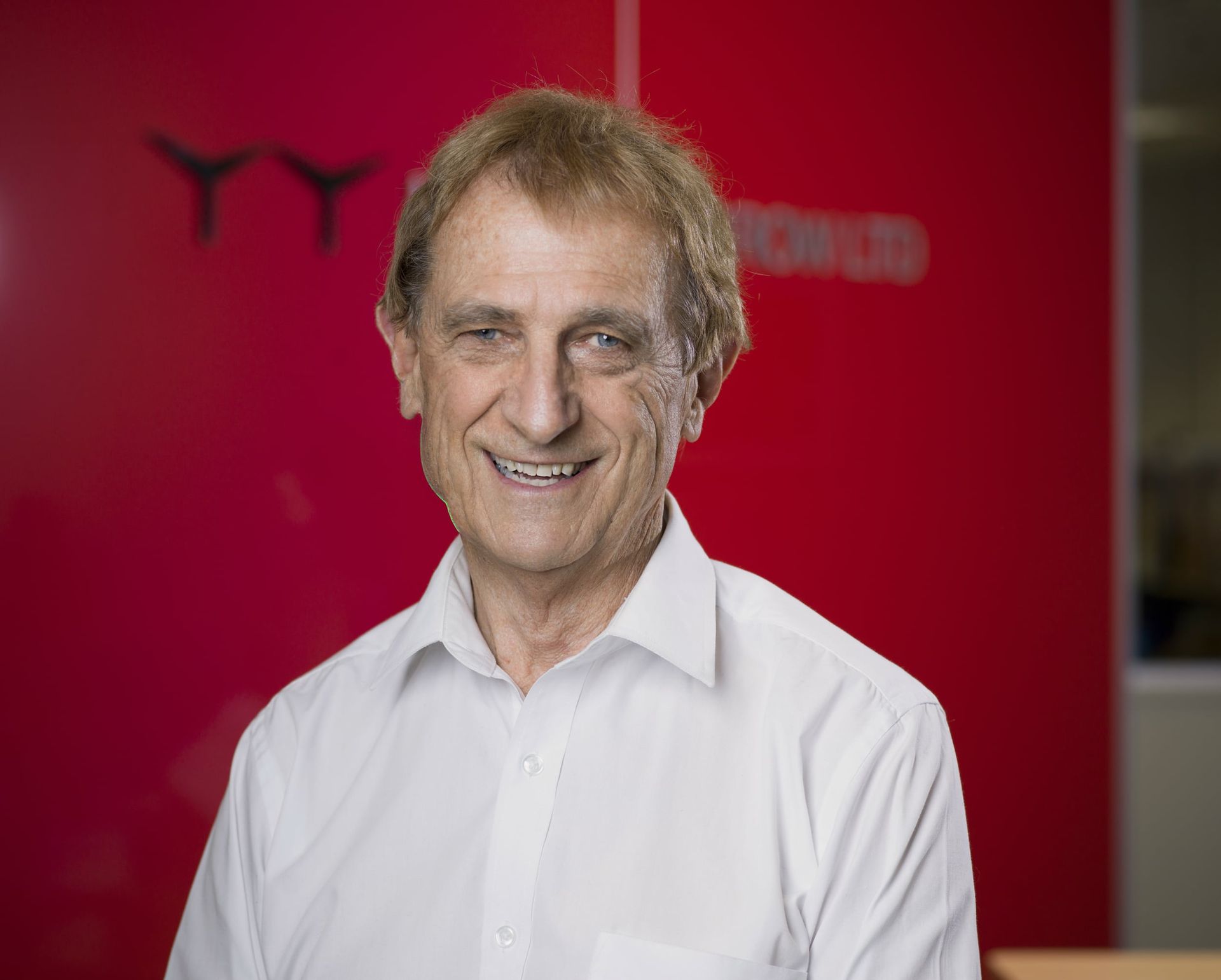 Man in white shirt smiling, standing in front of a red wall with a company logo.
