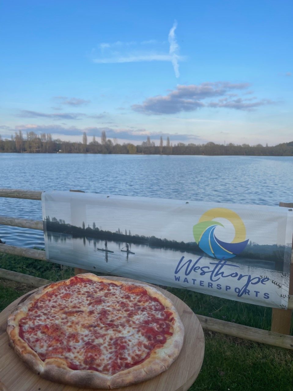 A pizza is sitting on a wooden table in front of a lake.