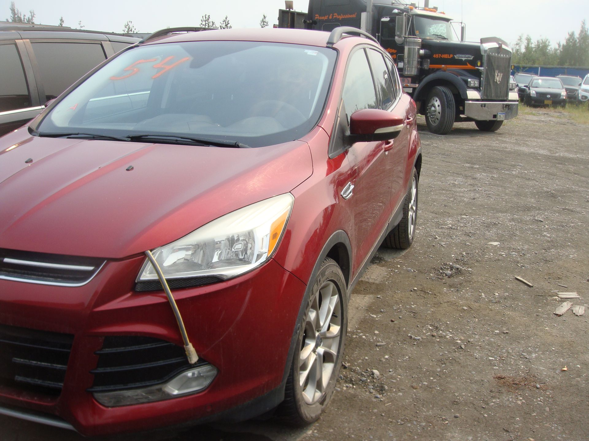 A red car is parked in a gravel lot next to a semi truck