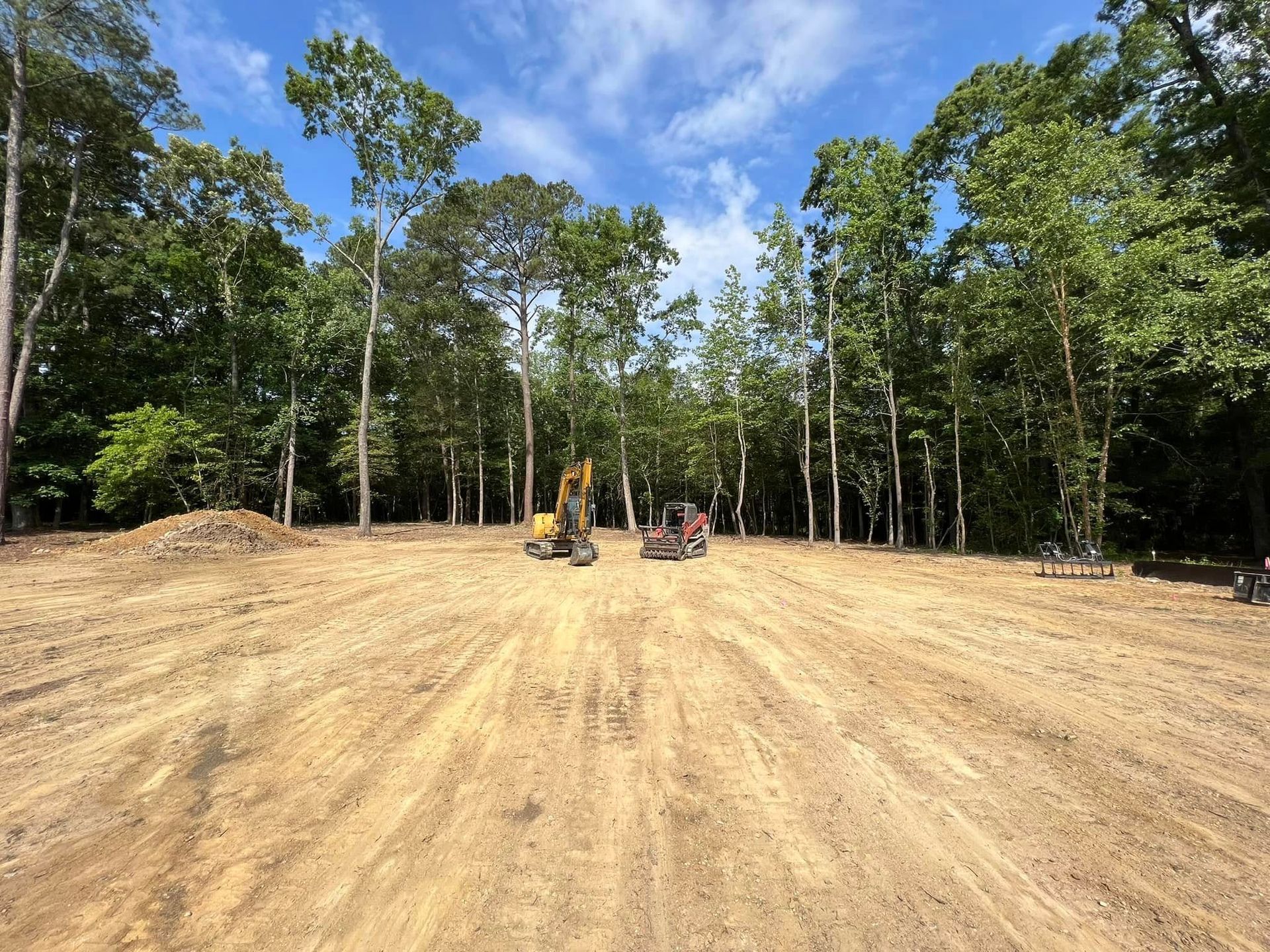 Heavy machinery sits on a cleared, graded dirt lot bordered by a forest of green trees under a bright blue sky.