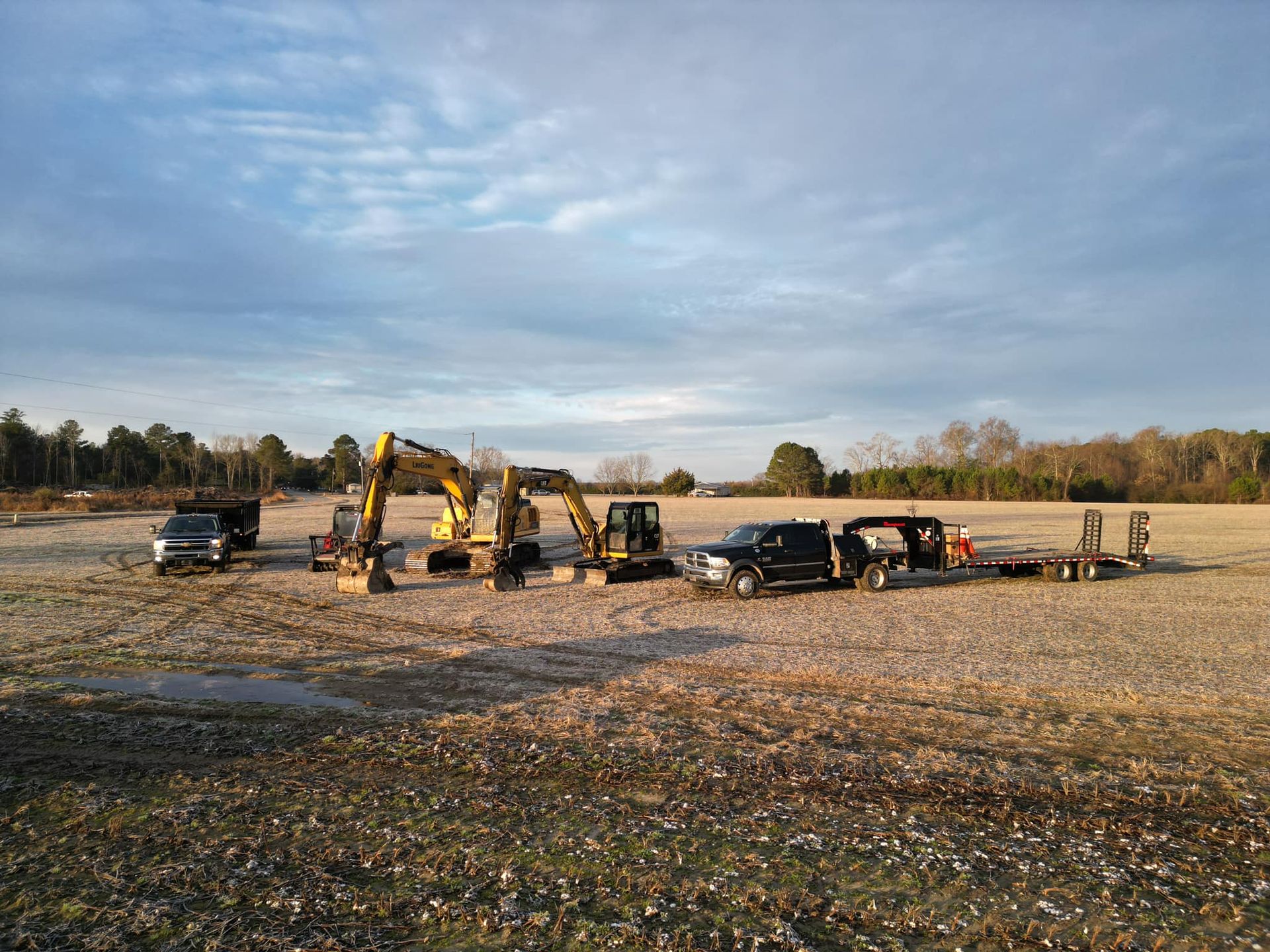 Yellow excavators and trucks parked on a large, open field under a blue sky at sunset.