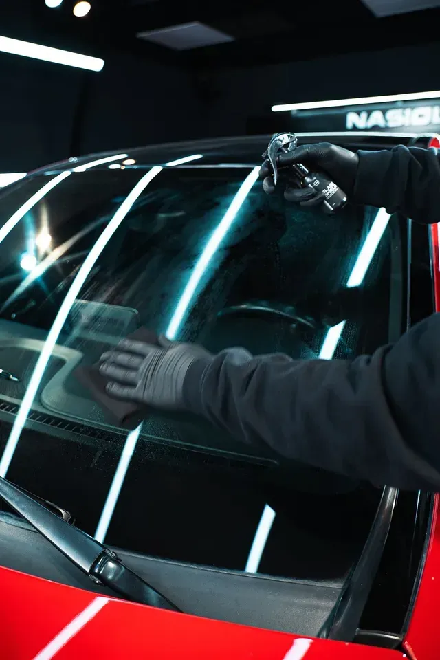 A person in black gloves cleans a car's windshield with a spray bottle and a microfiber cloth in a brightly lit studio.