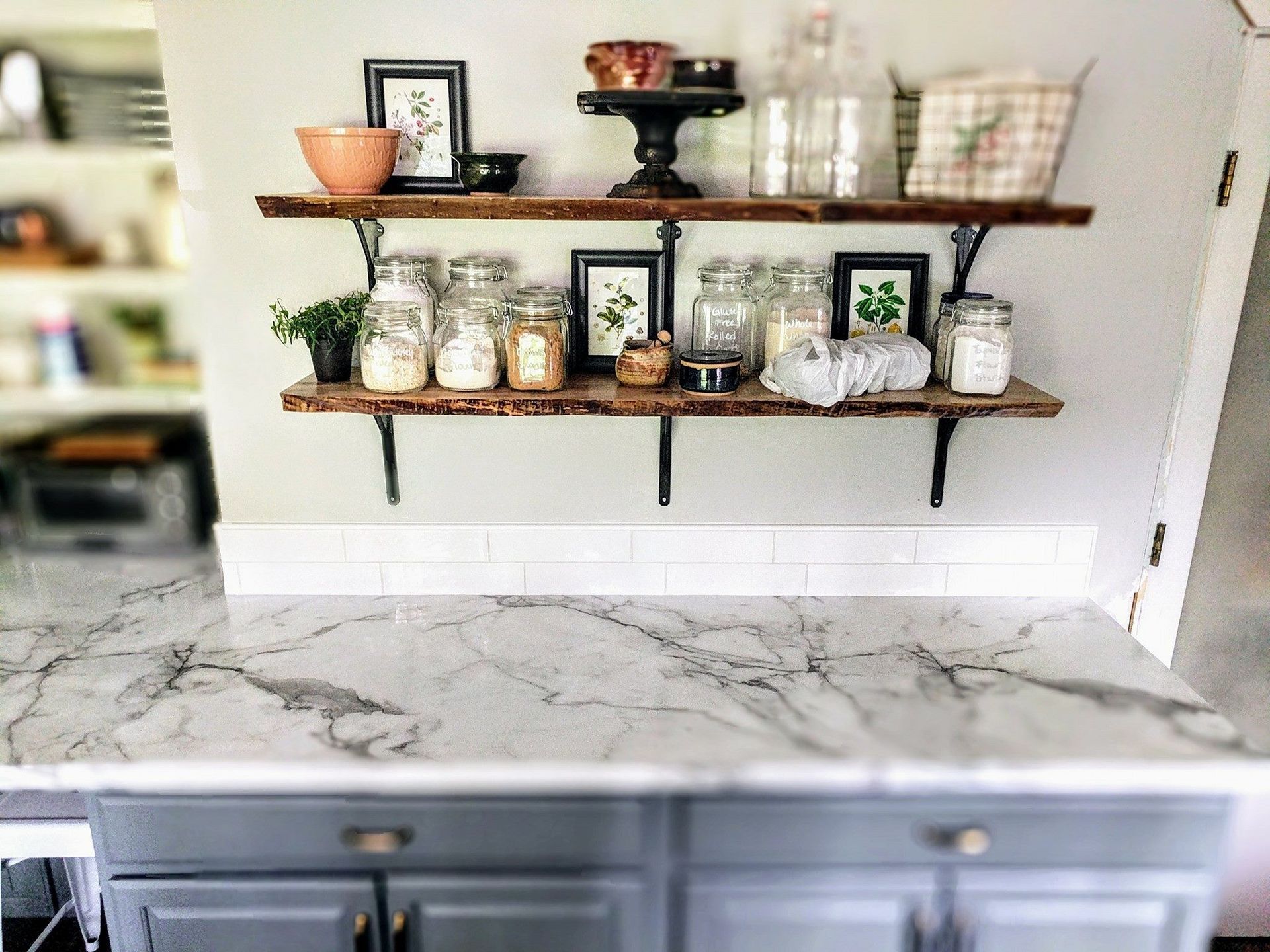 A kitchen with a marble counter top and wooden shelves filled with jars and pictures.