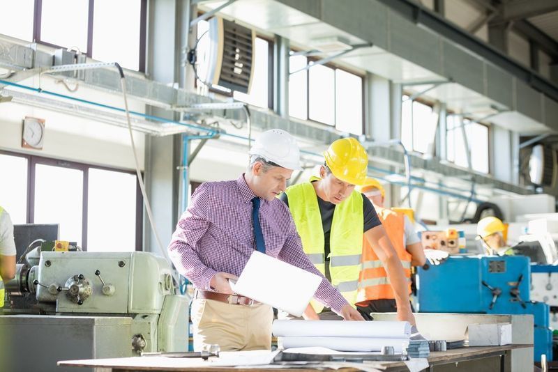 Two Men In Safety Gear Reviewing Blueprints In A Factory Setting — Wendy's Safety Services in Moulden, NT