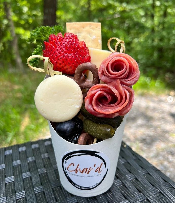 A cup filled with meat , cheese , strawberries and pretzels on a table.