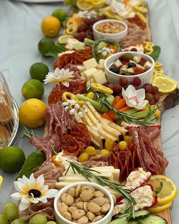 A long cutting board filled with meat , cheese , fruit , nuts and flowers.