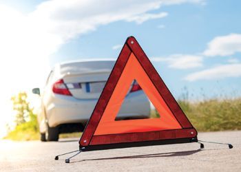 Red reflective triangle road hazard sign in front of a broken down white car on sunny road.