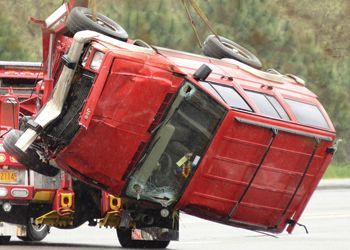 A red overturned truck being lifted by a tow truck; showing damage and debris at an outdoor scene.