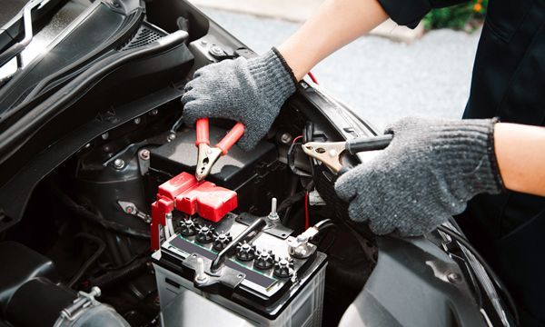 Hands in gray gloves connecting jumper cables to a car battery in an open engine bay.