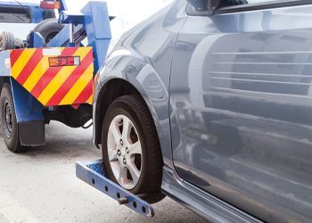 Tow truck towing a gray car, tire on a lift. Red and yellow warning on truck.