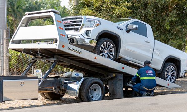 White GMC pickup truck being loaded onto a tow truck by a kneeling tow truck driver. Sunny outdoor setting.
