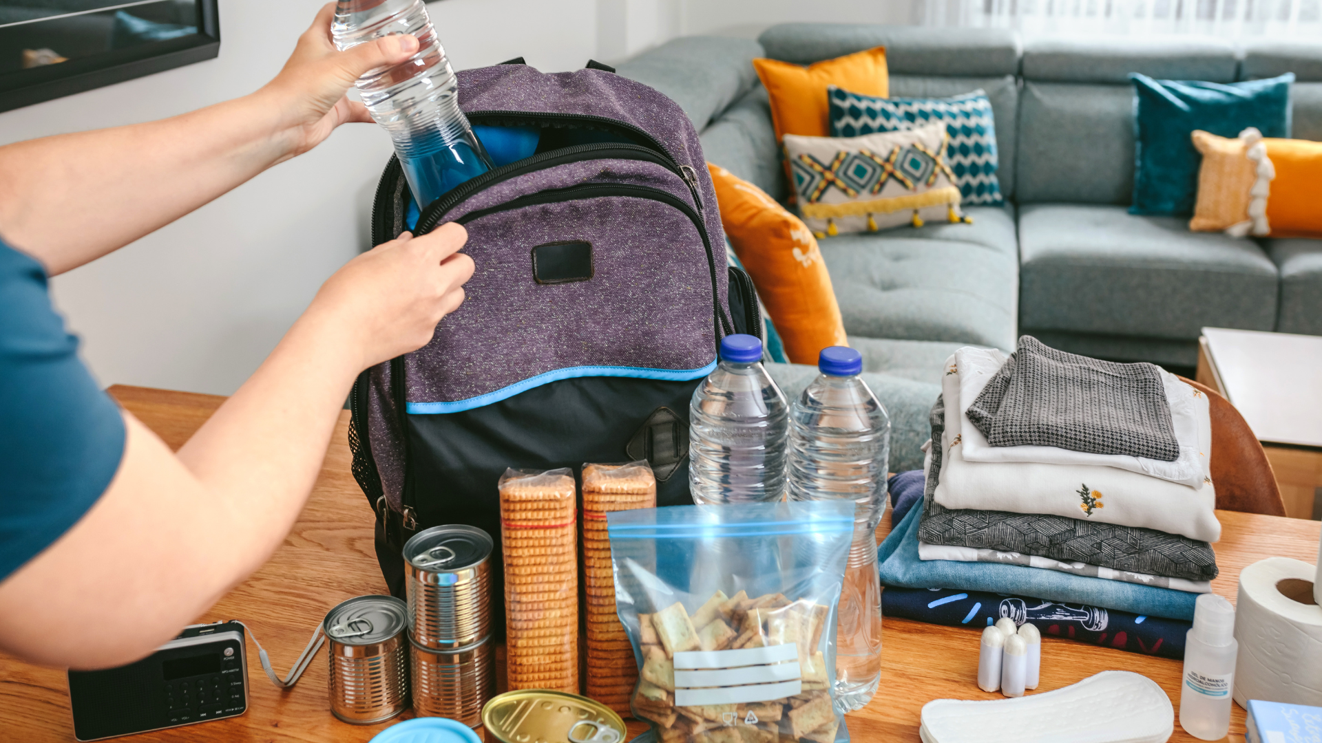 Person packing emergency supplies in a backpack on a table in a living room.
