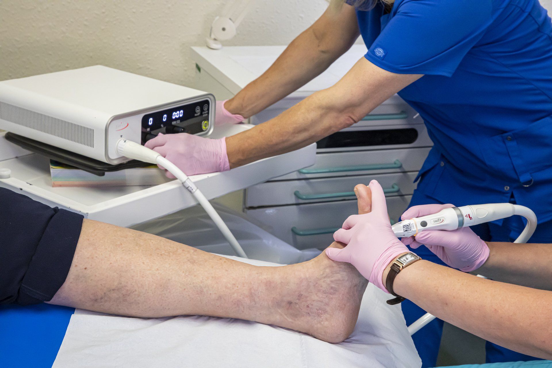 Medical professional uses device on a patient's foot. Hands wear gloves, device is white, patient is lying down.