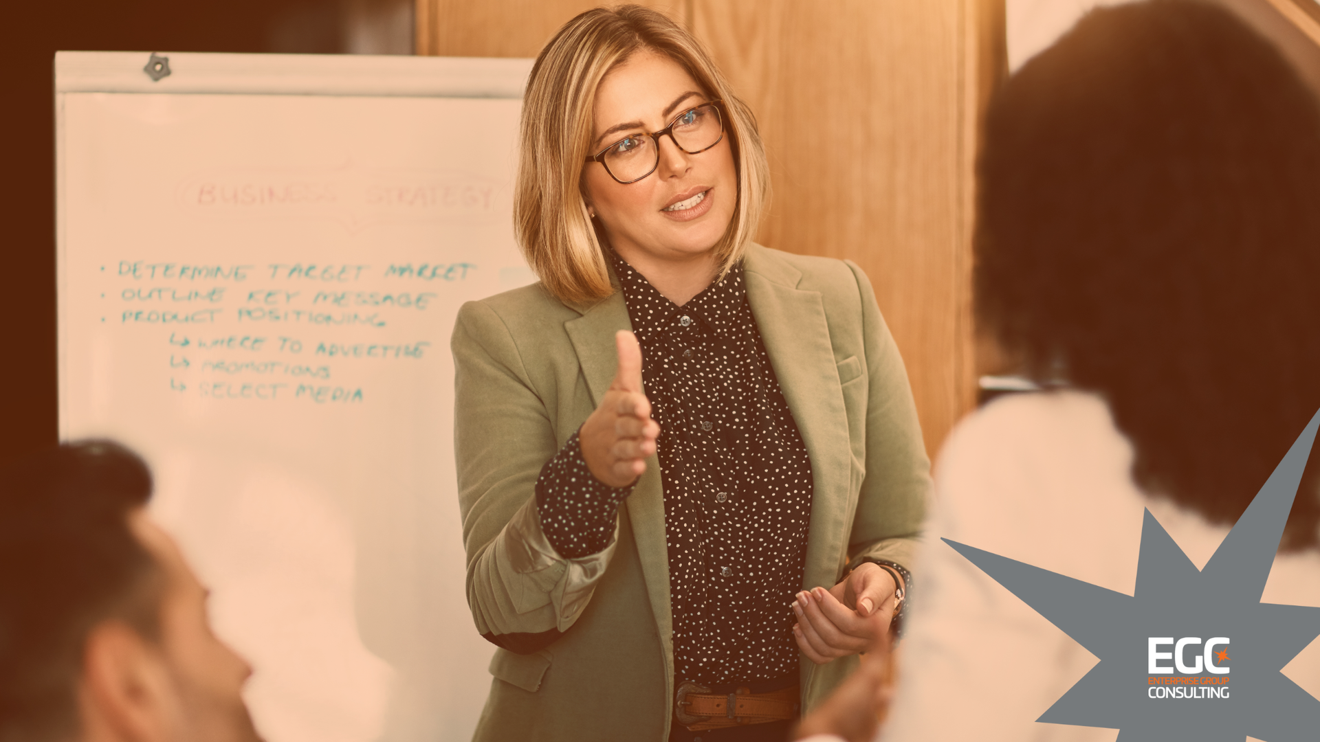 Woman in glasses giving a presentation, pointing at a whiteboard, talking to colleagues.