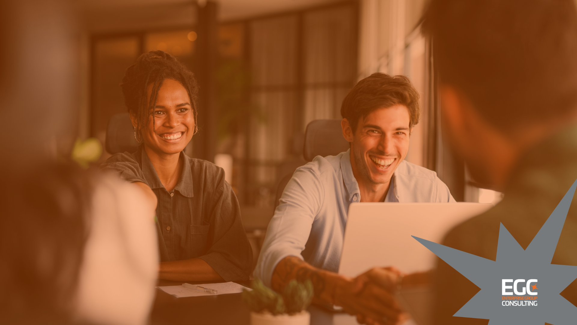 Two people smiling in a warm-toned office meeting, with a laptop and ELC logo in the corner.