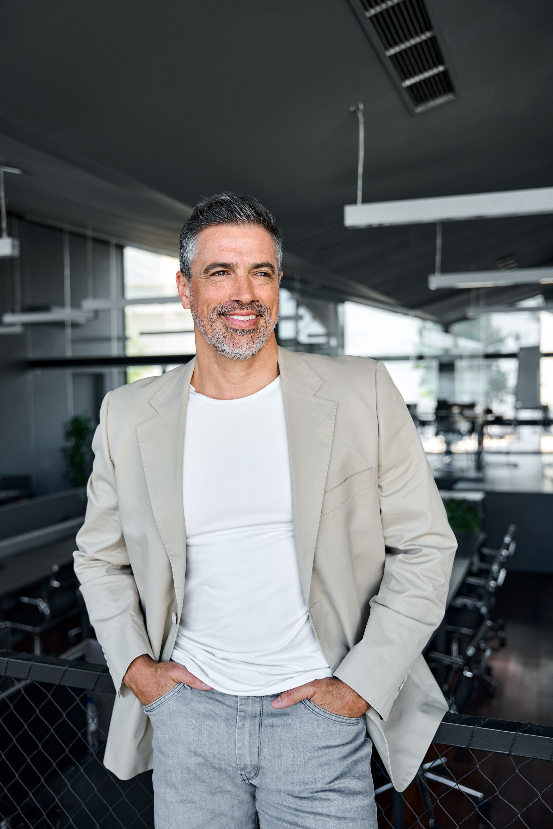 Man in blazer and jeans smiling in a modern office, hands in pockets.