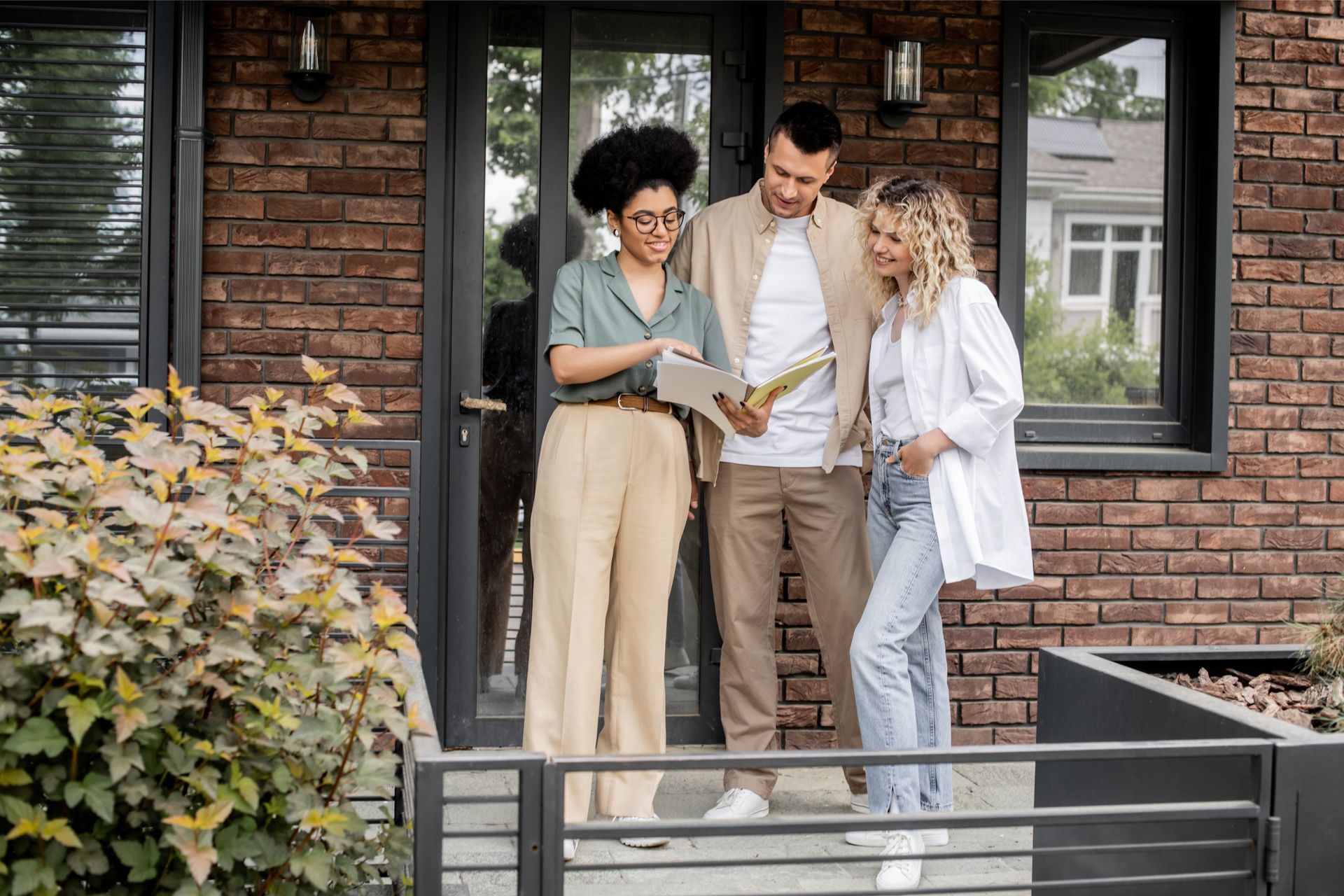 A realtor shows a document to a couple standing outside a brick house.