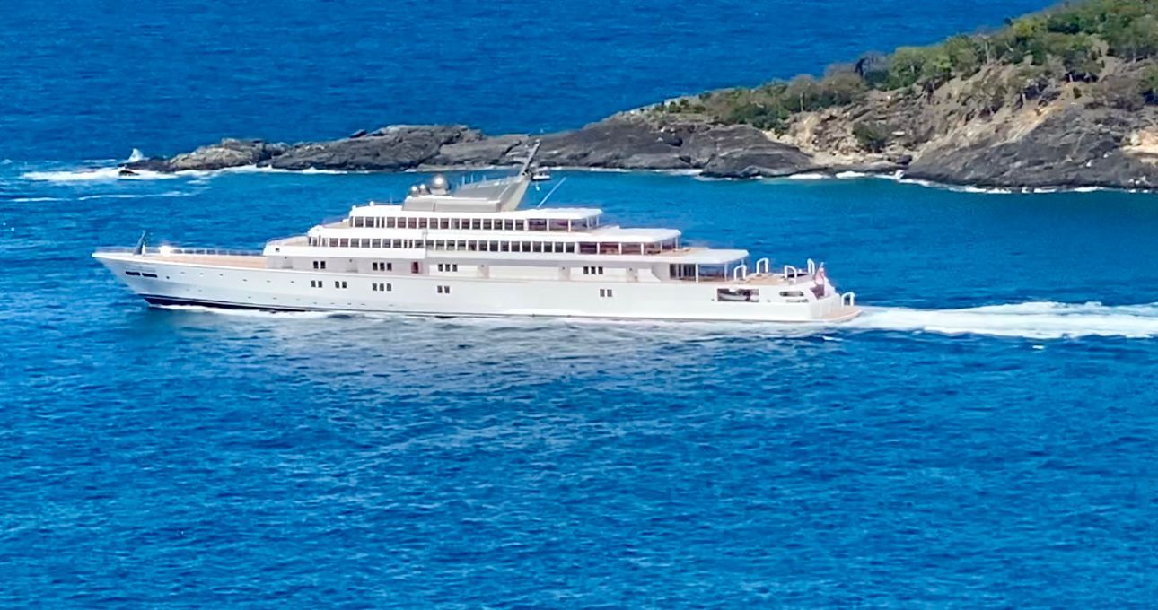 White multi-deck yacht sailing on blue water, with a rocky island in the background.
