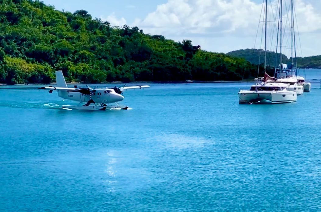 Seaplane landing in turquoise water near yachts, with lush green hills in the background.