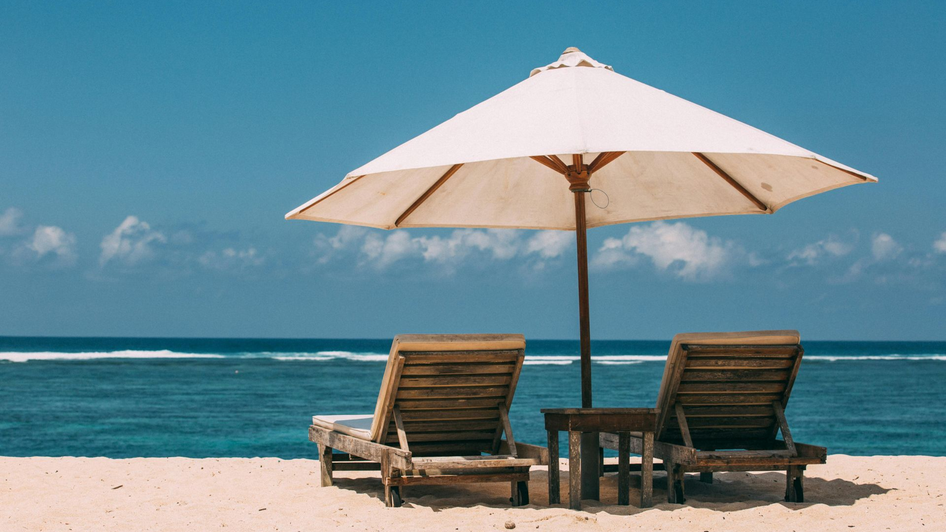 Two wooden beach chairs under a white umbrella on a sandy beach, facing the ocean under a blue sky.