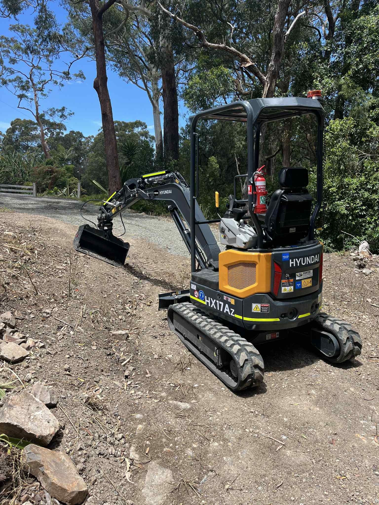 Small excavator on a gravel path near trees, under a blue sky.